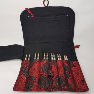 K&ouml;nnte beinhalten: Eine schwarze Stofftasche mit rotem Rosenmuster und einer Rei&szlig;verschlusstasche. Die Tasche ist ge&ouml;ffnet und enth&auml;lt ein Set mit 12 Stricknadeln.