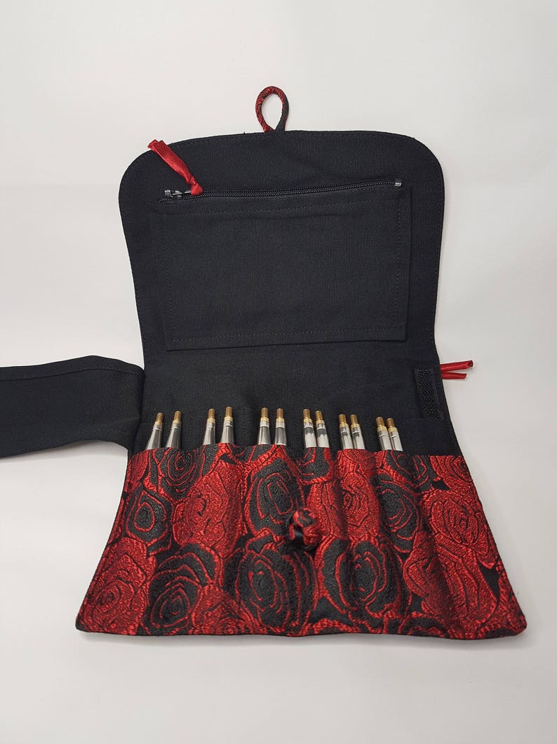 K&ouml;nnte beinhalten: Eine schwarze Stofftasche mit rotem Rosenmuster und einer Rei&szlig;verschlusstasche. Die Tasche ist ge&ouml;ffnet und enth&auml;lt ein Set mit 12 Stricknadeln.