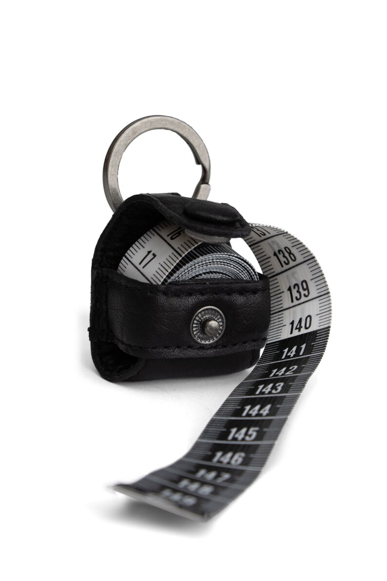 May include: A black leather keychain with a retractable tape measure. The tape measure is extended and shows numbers from 11 to 148.