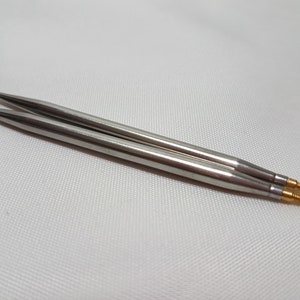 Puede incluir: Dos agujas de tejer plateadas con puntas doradas. Las agujas est&aacute;n sobre una superficie blanca.