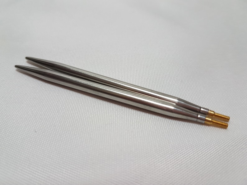 Puede incluir: Dos agujas de tejer plateadas con puntas doradas. Las agujas est&aacute;n sobre una superficie blanca.