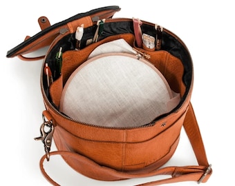 May include: A round, brown leather craft bag with a top zipper and a shoulder strap. The bag is open, revealing a linen embroidery hoop and various craft supplies, including needles and thread. The bag has a black interior and a small handle on the top.