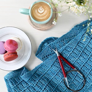 Könnte beinhalten: Ein blauer Strick Schal mit zwei roten Stricknadeln und einer Rundstricknadel. Es gibt auch Macarons und eine Tasse Kaffee auf einer weißen Oberfläche.