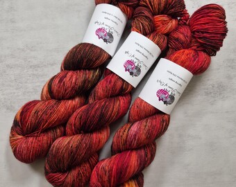 Könnte beinhalten: Drei Garnstränge in Fingering-Stärke mit einer bunten Farbpalette aus tiefem Rot, hellem Orange und schwarzen Sprenkeln. Jeder Strang ist mit einer weißen Papierbanderole versehen, die den Text "Dan's Flame of Craft" und "75% Merino 25% Nylon" trägt.
