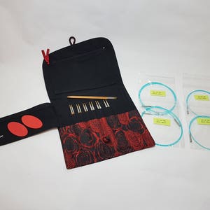 Puede incluir: Un estuche de tela negro y rojo con cierre de cremallera, que contiene un juego de agujas de tejer y un juego de agujas circulares. El estuche tiene un patr&oacute;n floral y un peque&ntilde;o bolsillo en la parte delantera. Las agujas est&aacute;n dispuestas en filas dentro del estuche.