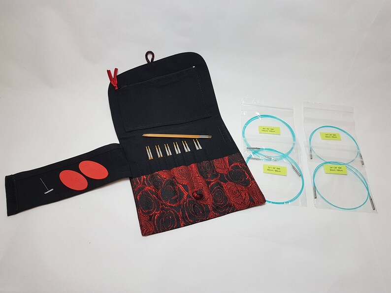 Puede incluir: Un estuche de tela negro y rojo con cierre de cremallera, que contiene un juego de agujas de tejer y un juego de agujas circulares. El estuche tiene un patr&oacute;n floral y un peque&ntilde;o bolsillo en la parte delantera. Las agujas est&aacute;n dispuestas en filas dentro del estuche.