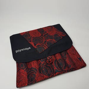 K&ouml;nnte beinhalten: Eine schwarze Stofftasche mit rotem und schwarzem Blumendesign. Die Tasche hat einen Knopfverschluss und das Wort "Hiyanhiya" ist auf der Vorderseite gestickt.