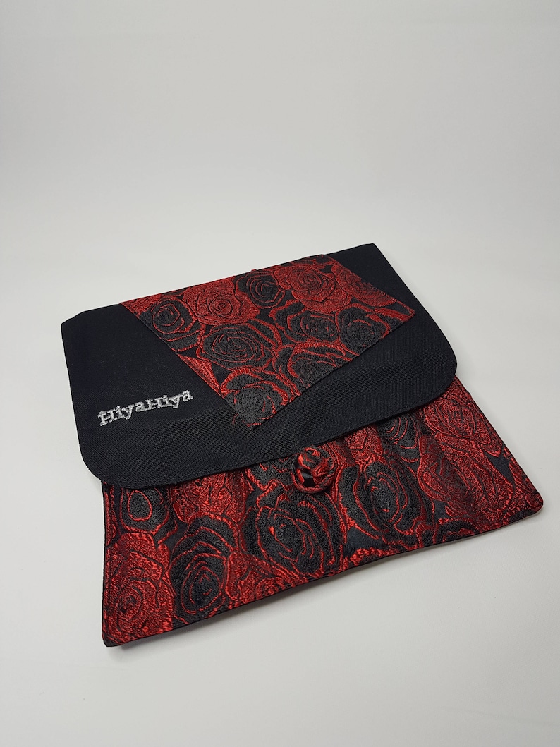 K&ouml;nnte beinhalten: Eine schwarze Stofftasche mit rotem und schwarzem Blumendesign. Die Tasche hat einen Knopfverschluss und das Wort "Hiyanhiya" ist auf der Vorderseite gestickt.