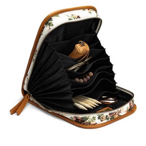 Può includere: Astuccio da lavoro a maglia con stampa floreale aperto con finiture e cerniera in pelle marrone. L'interno presenta più tasche nere contenenti ferri da maglia, un piccolo astuccio in pelle e altri accessori. L'astuccio è progettato per contenere forniture per maglieria.