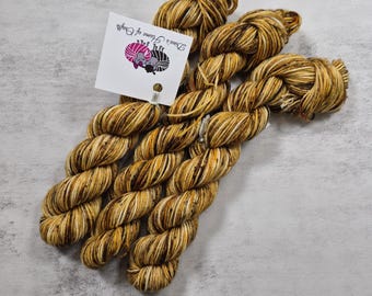 May include: Three skeins of hand-dyed yarn featuring a variegated color palette of golden yellow, deep brown, and cream white. A white square tag is attached to the yarn, displaying the text Dina's Home of Crafts and a logo of two sheep.