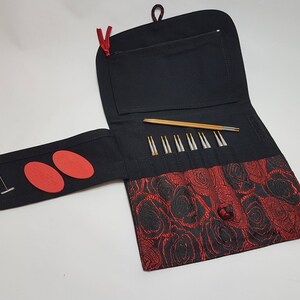 Puede incluir: Una funda de agujas de tejido negro con un patr&oacute;n de rosas rojas. La funda tiene una solapa que se abre para revelar un juego de agujas de tejer y un peque&ntilde;o bolsillo. Hay dos c&iacute;rculos de fieltro rojo en la solapa.