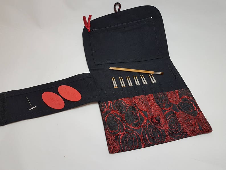 Puede incluir: Una funda de agujas de tejido negro con un patr&oacute;n de rosas rojas. La funda tiene una solapa que se abre para revelar un juego de agujas de tejer y un peque&ntilde;o bolsillo. Hay dos c&iacute;rculos de fieltro rojo en la solapa.