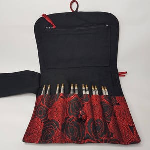 K&ouml;nnte beinhalten: Schwarze Nadeltasche mit rotem Rosenmuster und Rei&szlig;verschlussfach. Die Tasche ist ge&ouml;ffnet und zeigt einen Satz Stricknadeln.