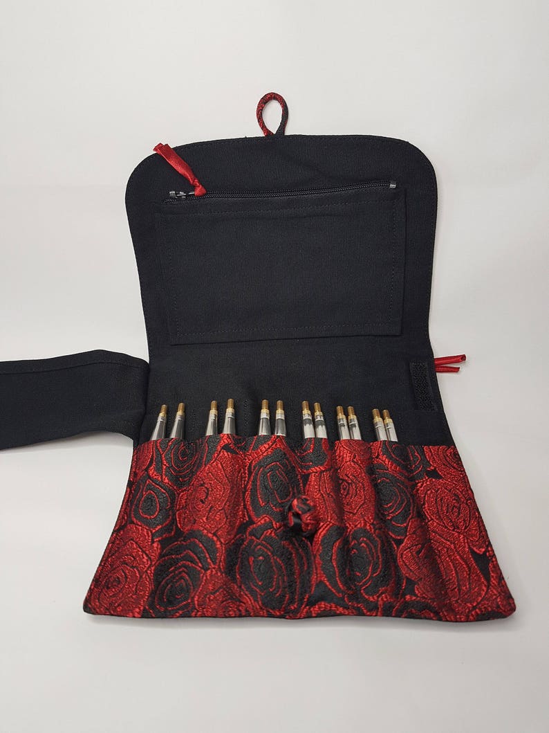 K&ouml;nnte beinhalten: Schwarze Nadeltasche mit rotem Rosenmuster und Rei&szlig;verschlussfach. Die Tasche ist ge&ouml;ffnet und zeigt einen Satz Stricknadeln.