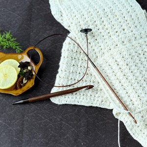 May include: Two wooden crochet hooks, one with a black plastic tip, on a white crocheted fabric. The fabric is on a dark brown surface.