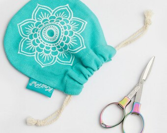 Puede incluir: Una bolsa de cordón turquesa con un diseño de mandala blanco y la palabra "KnitPro" impresa en la parte inferior. Un par de pequeñas tijeras de colores arcoíris está al lado de la bolsa.