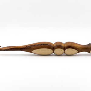 May include: A wooden crochet hook with a brown and light brown handle. The hook is labelled "Phoenix US L11/8.00mm".