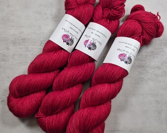 Könnte beinhalten: Drei Stränge leuchtend magentafarbenes Garn, jeweils mit einer weißen Papierbanderole versehen, die ein Logo mit einem Schaf und einem Garnknäuel sowie den Text 'Dye's Home of Craft' zeigt. Die Banderolen geben 70% Merino, 20% Seide und 10% Kaschmir an.