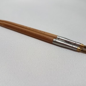 Può includere: Due ferri da maglia in legno con punte in metallo. Gli aghi sono realizzati in bambù marrone chiaro e hanno una finitura liscia e lucida. Le punte metalliche sono di colore argento e oro. Gli aghi sono progettati per lavorare a maglia e altri lavori artigianali.