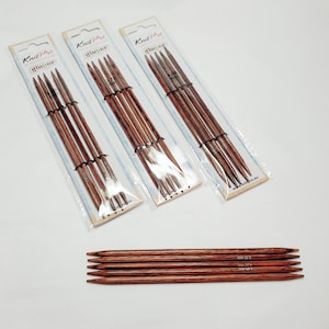 Puede incluir: Tres paquetes de agujas de tejer intercambiables Knit Pro Symfonie en tamaños 3,25 mm, 3,75 mm y 4,00 mm. Las agujas son de madera y tienen un color marrón rojizo. Los paquetes son blancos con detalles azules y rosas. Las agujas se muestran sobre una superficie blanca.