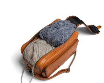 May include: A brown leather bag with a zippered top and a long strap. The bag is open and contains two balls of yarn and a gray knitted scarf.