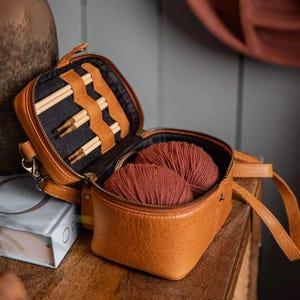 May include: A brown leather knitting bag with a zip closure and a strap. The bag is open and contains wooden knitting needles and a ball of yarn.