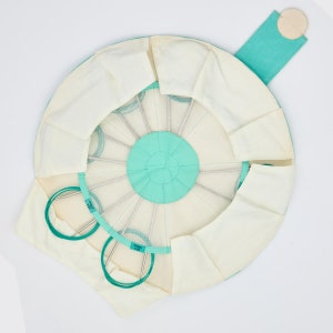 May include: A circular, teal-colored fabric case with a white interior. The case has multiple compartments for storing knitting needles. The case is open, revealing the needles inside. The needles are made of metal and have different sizes. The case has a small, white button on the side.