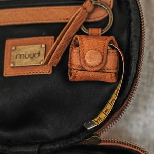 May include: A brown leather bag with a zipper closure and a small leather pouch attached to a key ring. The pouch has a button closure and the bag has a yellow measuring tape inside.