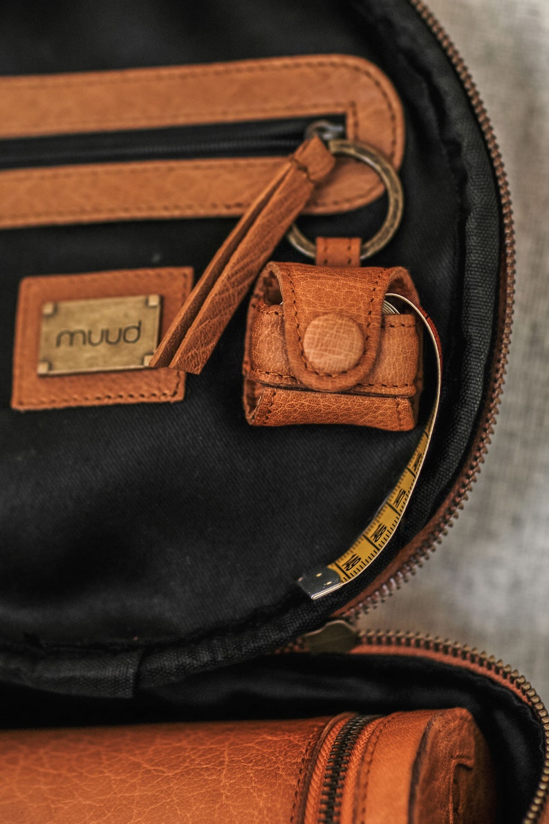 May include: A brown leather bag with a zipper closure and a small leather pouch attached to a key ring. The pouch has a button closure and the bag has a yellow measuring tape inside.