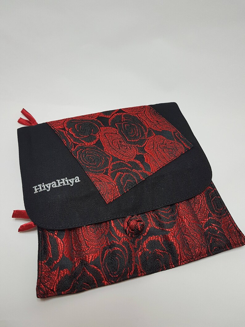 Puede incluir: Bolsa de tela negra con un patr&oacute;n de rosas rojas. La bolsa tiene una solapa con un patr&oacute;n de rosas rojas m&aacute;s peque&ntilde;o y el texto "HiyahHiya" bordado en blanco.