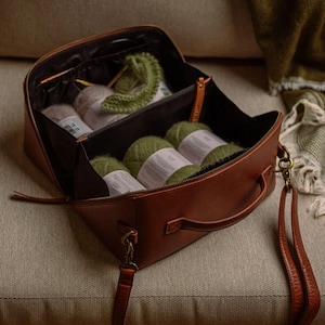 Può includere: Una borsa da lavoro a maglia in pelle marrone è aperta, rivelando diversi gomitoli di filato nei toni del verde e del beige. Sono visibili anche ferri da maglia e un piccolo campione lavorato a maglia. La borsa ha un manico e una tracolla staccabile.
