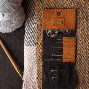 May include: A brown leather sewing needle case with a black fabric interior. The case has a small pocket with a metal label that says "muud". The case is open and filled with sewing needles and a small chain with beads.