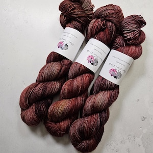 Puede incluir: Tres madejas de hilo teñido a mano en tonos burdeos, marrón y dorado. Cada madeja tiene una etiqueta blanca con texto y un logotipo. El hilo está dispuesto sobre una superficie de color claro, mostrando sus ricas variaciones de color y textura.