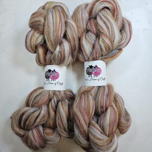 Può includere: Due gomitoli di lana cardata in un mix di colori marrone, bianco e beige. La lana cardata è disposta in un motivo intrecciato. I gomitoli sono etichettati con "Dina's Home of Crafts".