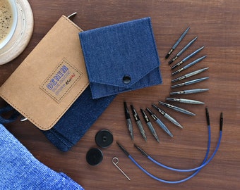 Puede incluir: Un juego de agujas de tejer intercambiables en una funda de transporte de temática vaquera. El juego incluye una variedad de tamaños de agujas, un cable y dos marcadores de puntos. La funda está hecha de tela vaquera azul con una solapa de cuero marrón que dice "DENIM SPECIAL COLLECTION EDITION Knit Pro".