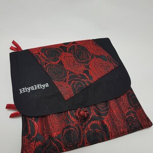 Puede incluir: Bolsa de tela negra con un patr&oacute;n de rosas rojas. La bolsa tiene una solapa con un patr&oacute;n de rosas rojas m&aacute;s peque&ntilde;o y el texto "HiyaHiya" bordado en blanco.