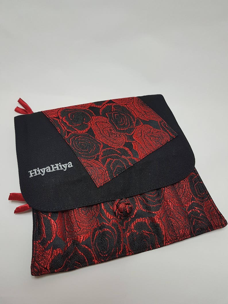 Puede incluir: Bolsa de tela negra con un patr&oacute;n de rosas rojas. La bolsa tiene una solapa con un patr&oacute;n de rosas rojas m&aacute;s peque&ntilde;o y el texto "HiyaHiya" bordado en blanco.