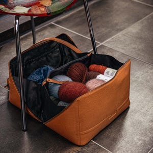 Può includere: Una borsa per filati in pelle marrone con una cerniera nera e un interno nero. La borsa è aperta e piena di gomitoli di filato di vari colori, tra cui blu, rosa, rosso e marrone.
