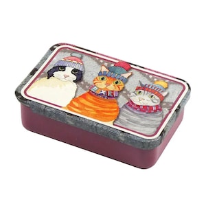 Puede incluir: Una caja de lata rectangular con tapa morada y base con estampado gris y blanco. La lata presenta un diseño de tres gatos con gorros de invierno y bufandas. Los gatos son un gato blanco y negro, un gato atigrado naranja y un gato atigrado gris.