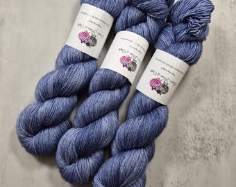 Könnte beinhalten: Drei Garnstränge in Fingering-Stärke in einer melierten blau-grauen Farbgebung. Jeder Strang hat ein weißes Papieretikett mit dem Text "Dye's Flame of Craft" und einem Logo aus zwei Schafen, das eine Mischung aus 70% Merino, 20% Seide und 10% Kaschmir angibt.