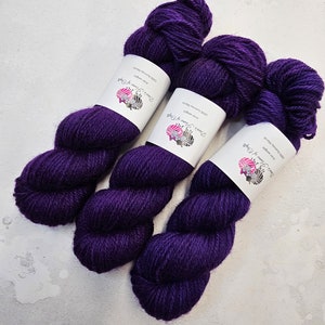 Könnte beinhalten: Drei Knäuel tiefes violettes Garn, jeweils mit dem Etikett "Dina's Flame of Yarn" und dem Text "100% Romney Marsh Aran Weight".