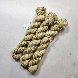 May include: Three skeins of yarn in a green and cream colorway with brown speckles. The yarn is twisted into a rope shape and is ready to be used for knitting or crocheting.