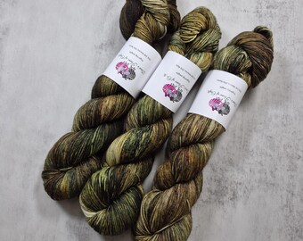 Könnte beinhalten: Drei Garnstränge in Fingering-Stärke mit einer melierten Farbpalette aus Dunkelbraun, Olivgrün und goldenen Akzenten. Jeder Strang ist mit einer weißen Papierbanderole versehen, die den Text "Dye & Flame of Craft" sowie Faserangaben enthält.