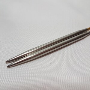 Puede incluir: Dos agujas de tejer plateadas con puntas doradas. Las agujas est&aacute;n sobre una superficie blanca.