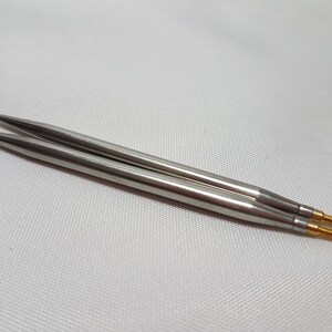 Puede incluir: Un par de agujas de tejer plateadas con puntas doradas. Las agujas est&aacute;n sobre una superficie blanca.