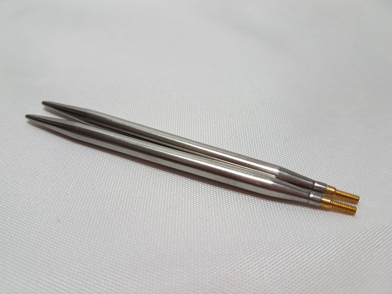 Puede incluir: Un par de agujas de tejer plateadas con puntas doradas. Las agujas est&aacute;n sobre una superficie blanca.