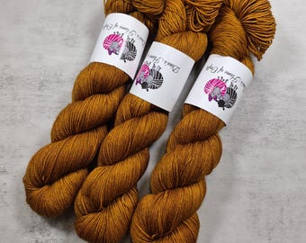 Könnte beinhalten: Drei Garnstränge in Goldbraun, jeweils umwickelt mit einem weißen Papieretikett, das ein Logo eines stilisierten Schafs mit rosa und schwarzen Akzenten sowie den Text Dina's Home of Crafts zeigt. Das Garn hat eine gedrehte Struktur.