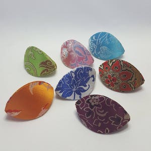 May include: Eight fabric hair clips in various colours and patterns. The clips are shaped like a half-moon and have a floral design. The colours include green, pink, blue, orange, red, white, and purple.