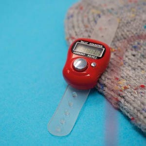 Peut inclure: Un compteur numérique rouge avec un bracelet transparent. Le compteur a des boutons marqués "Compter" et "Réinitialiser". Le compteur est sur une surface bleue à côté d'un tissu tricoté gris avec des points colorés.