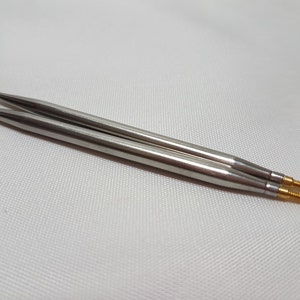 Puede incluir: Dos agujas de tejer plateadas con puntas doradas. Las agujas est&aacute;n sobre una superficie blanca.